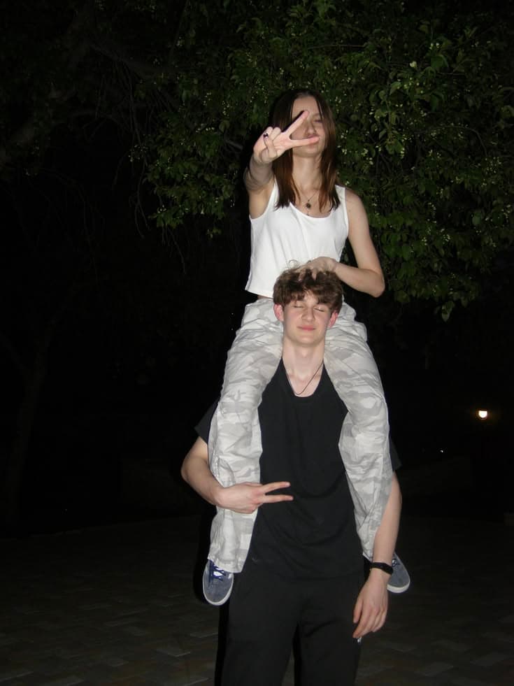 A girl sitting on a boy's shoulders at night, both making peace signs